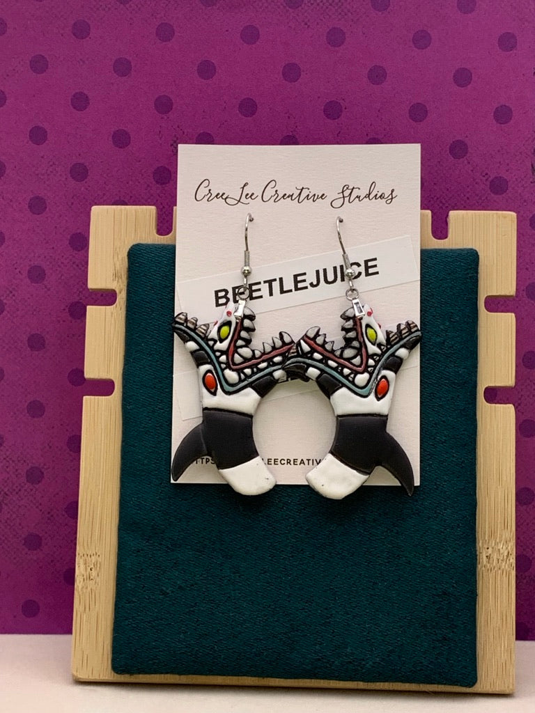 A pair of Beetlejuice-inspired sandworm character earrings displayed on a card, with the earrings featuring black-and-white stripes and vibrant red, yellow, and green patterns.
