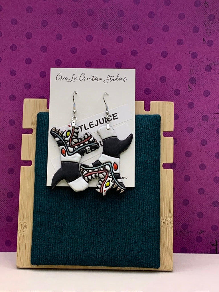 A pair of handcrafted polymer clay earrings inspired by the movie Beetlejuice, displayed on a stand with a purple background.
