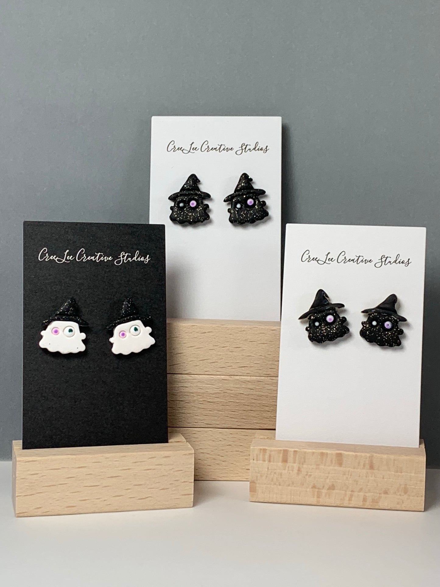 A white ghost with sparkle black hat stud earring and sparkle black ghosts with sparkle black hats stud earrings on a business card in a wooden stand.