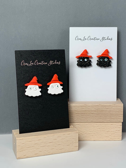 A white ghost with orange hat stud earring and a black ghost with orange hat stud earring on a business card in a wooden stand.