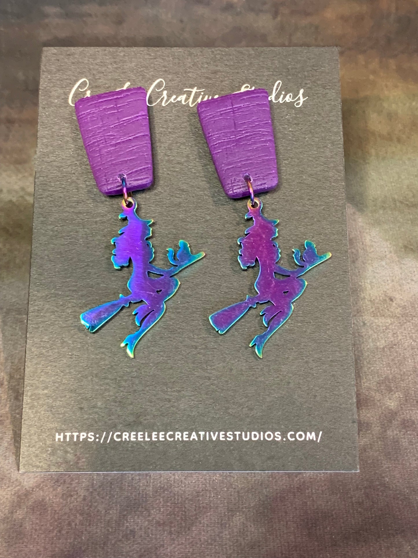A pair of vibrant purple dangle earrings with a stainless steel witch charm.