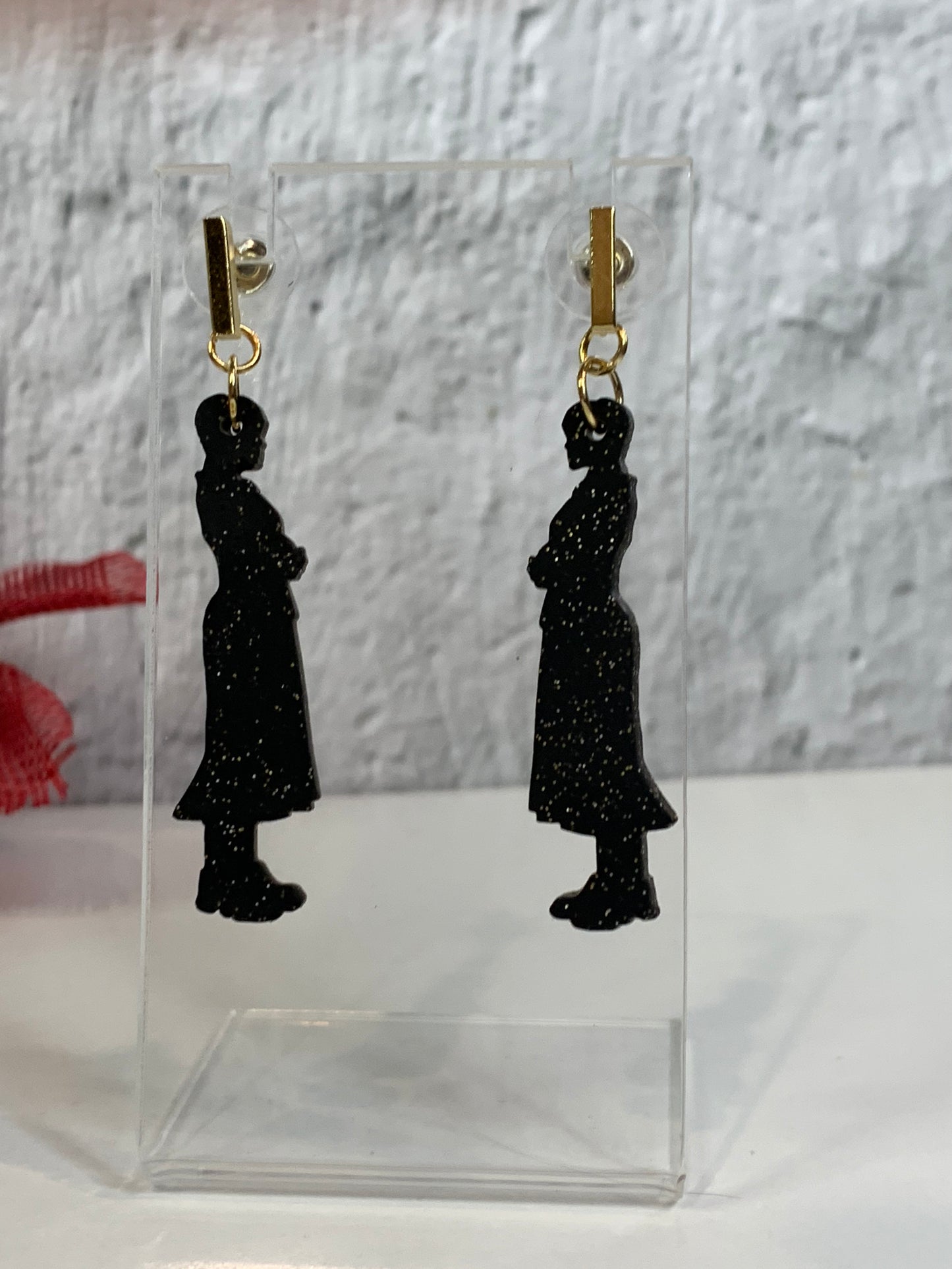 Black silhouette earrings of Wednesday Addams on clear stands against a gray background