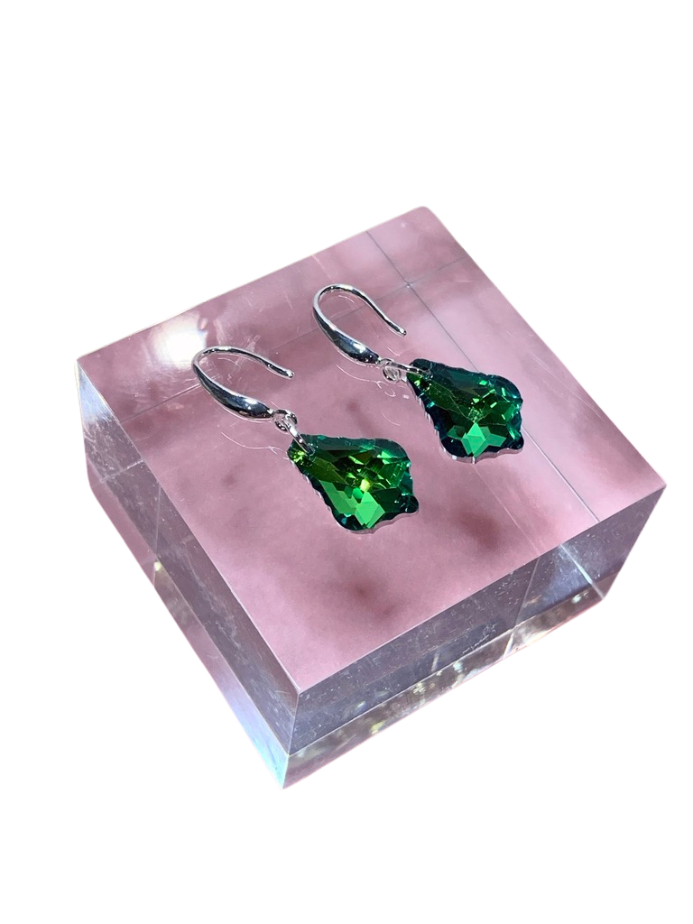A pair of vibrant green crystal earrings displayed on a pink stand, with a reflective surface in the background.