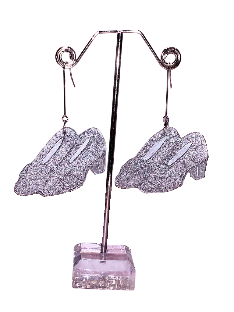 A pair of silver glittery slipper earrings displayed on a stand with a Wicked-themed design, reminiscent of Elphaba’s sister’s slippers and Cinderella’s shoes.
