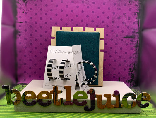 A pair of acrylic black and white striped hoop earrings displayed on a stand with a 'Beetlejuice' text sign in the background, all against a purple dotted background.