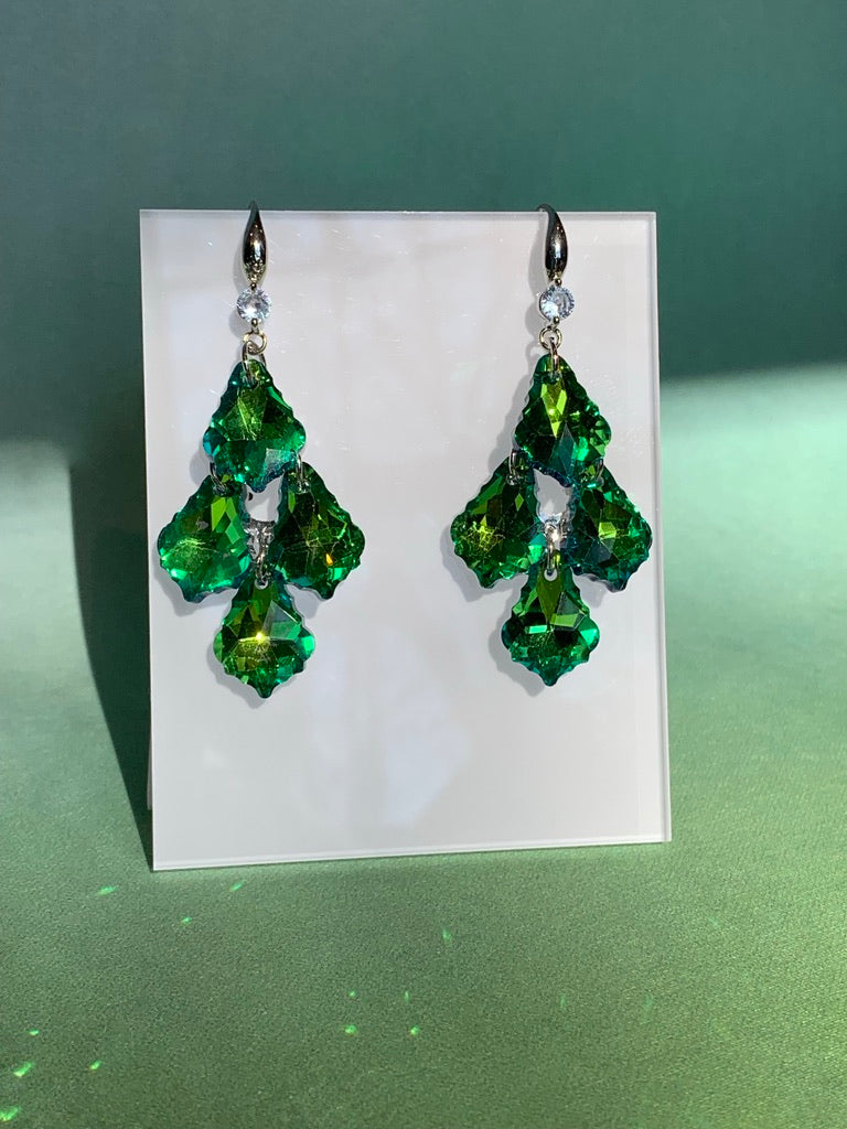 A pair of emerald green tiered palm crystal dangle earrings displayed on a white stand with a green background.