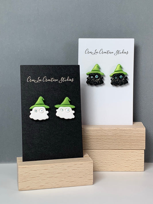 A white ghost with green hat stud earring and a black ghost with green hat stud earring on a business card in a wooden stand.