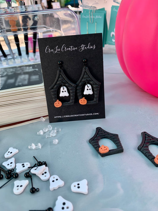 Halloween themed earrings shaped like a haunted house with a spooky sheet ghost floating inside. Background is a table with components of the earrings around ready to be assembled. 