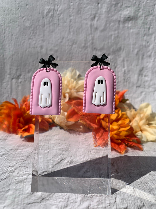 Pink ghost earrings with black bows on a clear stand against a textured gray background.