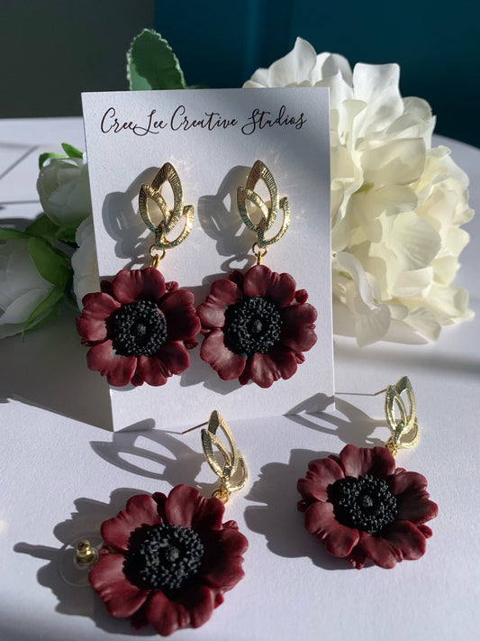 A pair of maroon poppy flower earrings with gold-colored stems and leaves, displayed on a white card with a floral background.