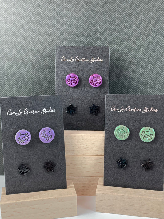 Three pairs of stud earrings displayed on black cardboards. Each pair features a detailed design of a spider or a spiderweb, with colors including purple, magenta (middle) and green.