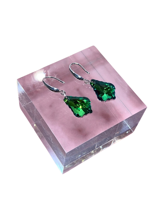 A pair of vibrant green crystal earrings displayed on a pink stand, with a reflective surface in the background.