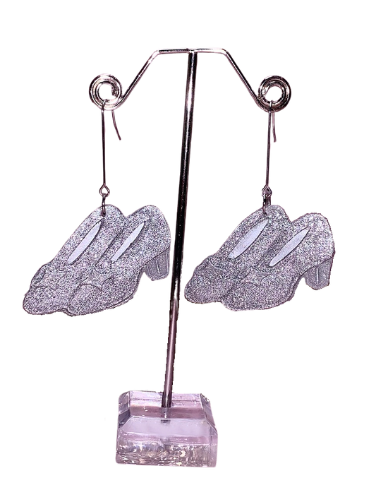 A pair of silver glittery slipper earrings displayed on a stand with a Wicked-themed design, reminiscent of Elphaba’s sister’s slippers and Cinderella’s shoes.